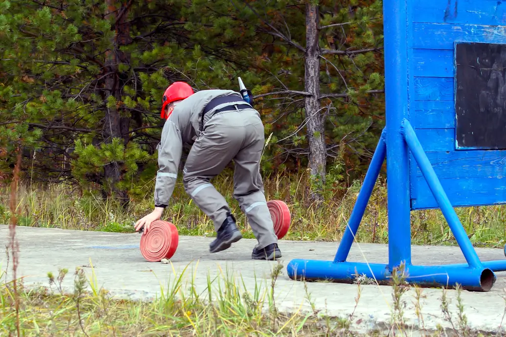 Candidat pompier en uniforme et casque rouge manipulant des tuyaux lors d'un test physique d'aptitude en extérieur.