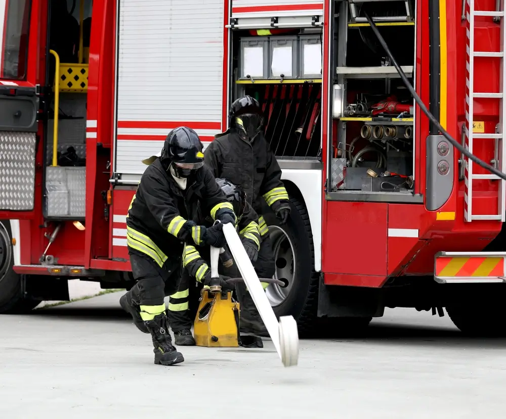 Pompiers en formation manipulant une lance et un tuyau d'incendie devant un camion de secours lors d'un exercice.
