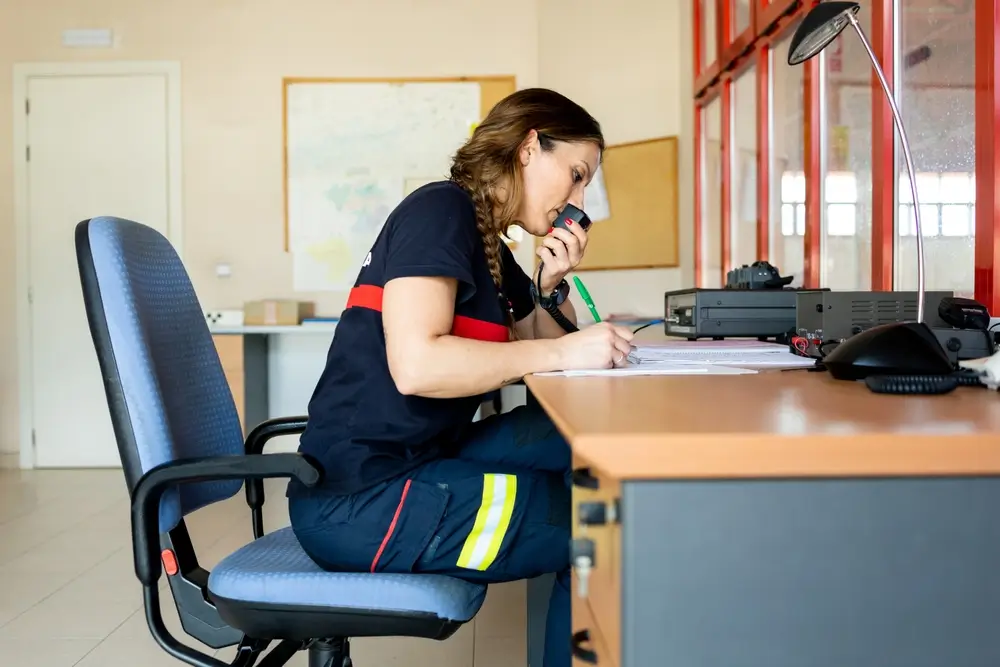 Sapeur-pompier en uniforme dans un centre d'appel d'urgence, communiquant par radio et prenant des notes à son bureau.