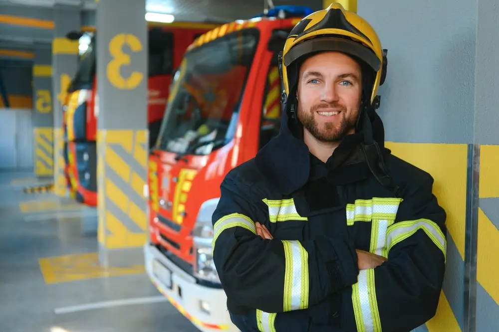 Sapeur-pompier professionnel en uniforme de feu et casque, posant devant des camions rouges dans son centre de secours.
