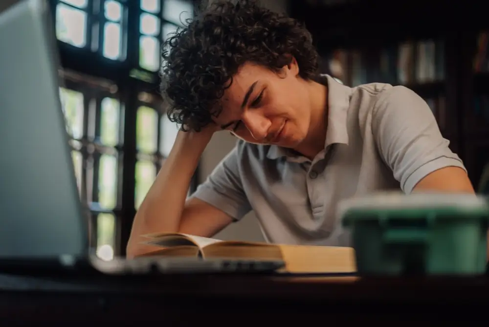 Jeune homme concentré révisant ses cours avec un manuel de préparation au concours devant son ordinateur.