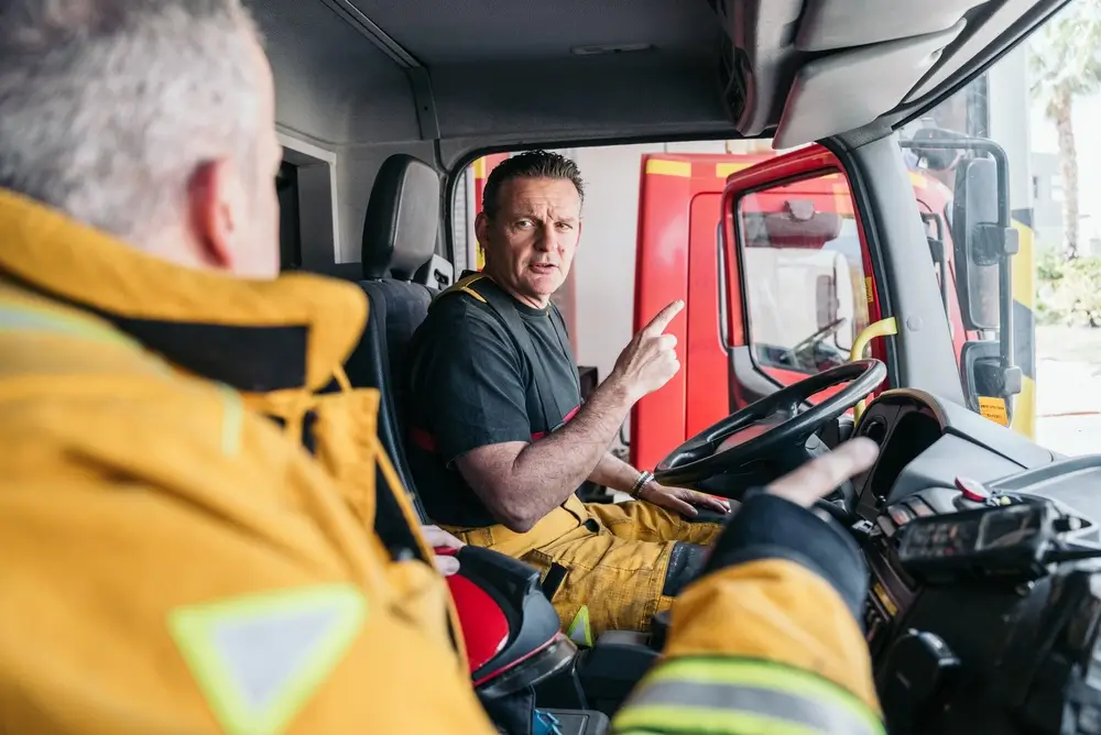 Pompiers en tenue échangeant dans la cabine d'un camion d'incendie, un casque de pompier posé sur le tableau de bord.