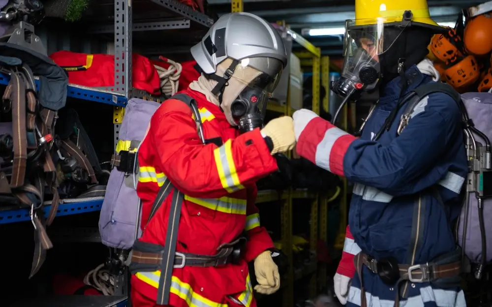 Deux pompiers en tenue complète avec casque de pompier se saluent dans un local de rangement d'équipement.