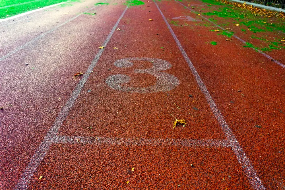 Piste d'athlétisme rouge avec des lignes blanches et le numéro 3 au sol pour une épreuve de course d'endurance.