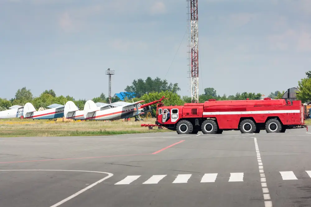 Camion de secours incendie rouge stationné sur le tarmac d'un aérodrome avec des avions légers en arrière-plan.